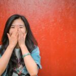 Young overjoyed Asian female in casual outfit covering face with hands and laughing while standing against vibrant orange wall